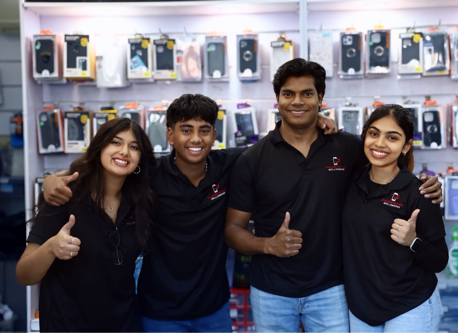 The Phone Repair Wollongong team in black branded polo shirts, giving a thumbs up in front of the shop’s wall of phone cases.